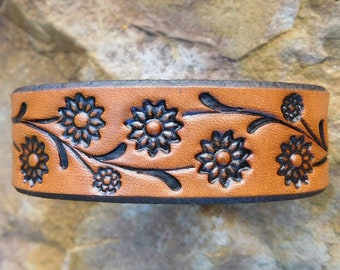 May include: A brown leather cuff bracelet with a floral design. The bracelet features embossed black flowers and leaves. The edges are dark brown, and the overall design is rustic and handcrafted.