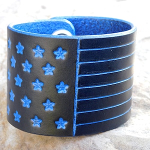 May include: A black leather bracelet with a blue outline and a design of the American flag. The stars are embossed and the stripes are stitched.