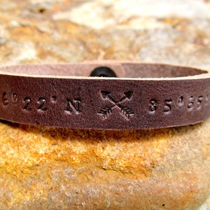 May include: A brown leather bracelet with the coordinates "36&deg;22' N 85&deg;65'" and two crossed arrows engraved on it.
