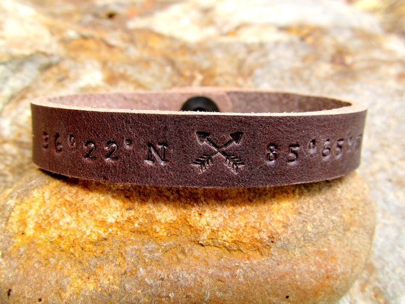 May include: A brown leather bracelet with the coordinates "36&deg;22' N 85&deg;65'" and two crossed arrows engraved on it.