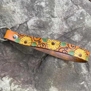 May include: A brown leather keychain with a sunflower design. The keychain has a silver metal ring and is decorated with yellow sunflowers and green leaves.