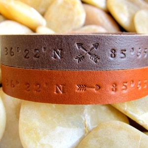 May include: A brown and orange leather bracelet with coordinates and arrows engraved on it. The coordinates are 36&deg; 22' N 85&deg; 65' W.