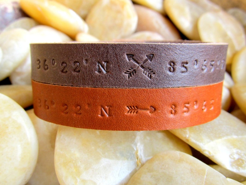 May include: A brown and orange leather bracelet with coordinates and arrows engraved on it. The coordinates are 36&deg; 22' N 85&deg; 65' W.