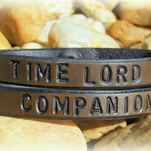 May include: Two brown leather bracelets with black stitching. The top bracelet has the words "TIME LORD" embossed on it. The bottom bracelet has the words "COMPANION" embossed on it.