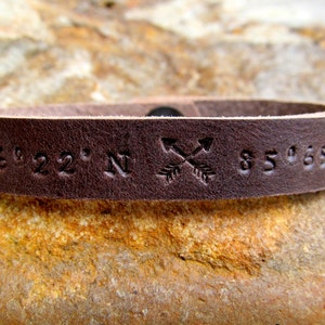 May include: A brown leather bracelet with the coordinates "36&deg;22' N 85&deg;65' W" and two crossed arrows engraved on it.