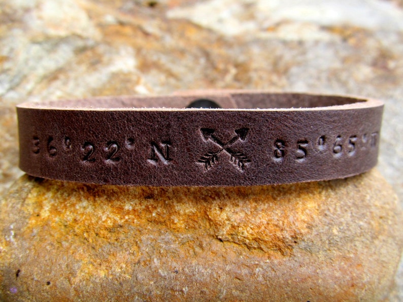 May include: A brown leather bracelet with the coordinates "36&deg;22' N 85&deg;65' W" and two crossed arrows engraved on it.