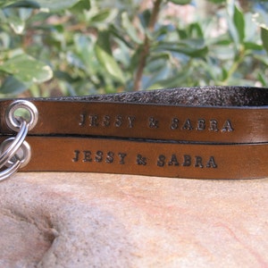 May include: Two brown leather keychains with silver key rings. The keychains are engraved with the text "Jessy & Sabra".