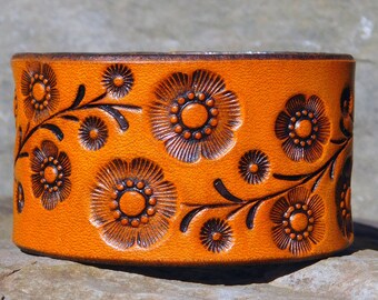May include: A brown leather cuff bracelet with a floral design embossed into the leather. The flowers are brown and have a raised center.