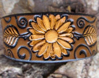 May include: A brown leather bracelet with a tooled design of a daisy flower and swirling vines. The flower is in shades of brown and yellow.