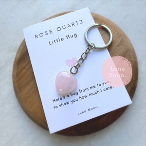 May include: A pink rose quartz heart-shaped keyring with a silver chain. The keyring is presented on a white card with the text "ROSE QUARTZ Little Hug" and a message. The background is a wooden surface.