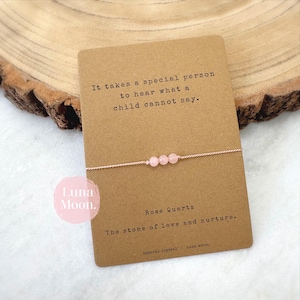 May include: A rose quartz bracelet with three pink beads on a fine cord. The bracelet is presented on a brown card with the text "It takes a special person to hear what a child cannot say."