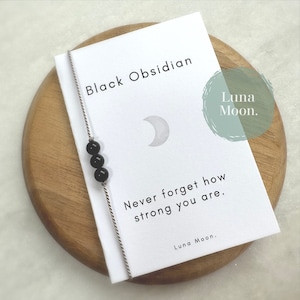 May include: A white card with the text "Black Obsidian" and a crescent moon graphic. The card also includes the text "Never forget how strong you are." and "Luna Moon." A black obsidian beaded bracelet is attached to the card.