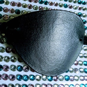 Könnte beinhalten: Eine schwarze Augenklappe mit einem elastischen Band. Die Augenklappe besteht aus einem strukturierten Material und ist für ein Auge konzipiert. Der Hintergrund ist mit bunten Strasssteinen verziert.