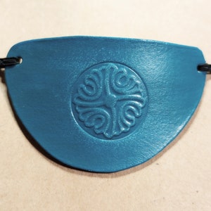 Op de afbeelding: Teal ooglapje met een decoratief, cirkelvormig reliëfontwerp. Het ooglapje is gemaakt van een glad materiaal en heeft een zwart koord om het vast te zetten. Het ontwerp is een kruisvormig patroon.