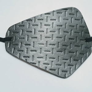 May include: A black eye patch with a textured, diamond-plate pattern. The patch is connected to a black elastic band. The eye patch is designed to cover one eye.