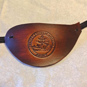 Peut inclure: Un cache-œil en cuir marron avec un emblème de l'armée américaine en relief. Le cache-œil a une forme incurvée et une sangle élastique pour un port sûr. L'emblème présente un design circulaire avec du texte et une illustration détaillée.