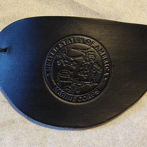 May include: Black leather eye patch with an embossed United States Marine Corps emblem. The emblem is circular and features an eagle, globe, and anchor design, with text reading "United States of America" and "Marine Corps". An elastic strap is attached.