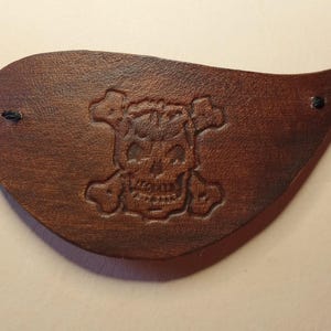 Könnte beinhalten: Ein braunes Leder-Piraten-Augenpflaster mit einem Totenkopf- und Knochenkreuz-Design.