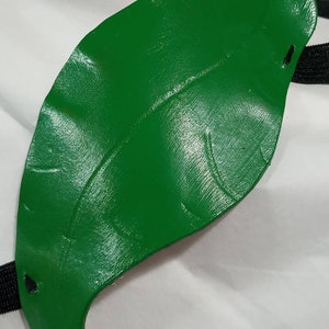 May include: A green leather eye patch with a black elastic strap. The patch is shaped like a leaf and has a textured surface.