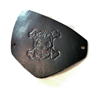 May include: A black leather patch with a shield-like shape featuring an embossed skull and crossbones design in the centre. The patch has a smooth, dark surface and includes two small circular holes, one on each side for attachment purposes.
