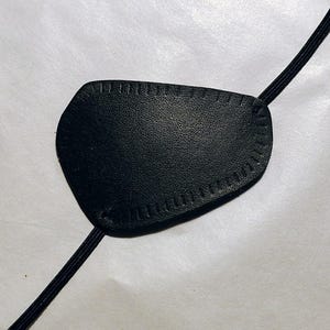 May include: Black eye patch with a textured surface and stitched edges. The patch is connected to a black elastic band. The eye patch is designed to cover one eye.