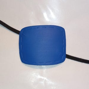 May include: A bright blue eye patch with a stitched border and a black elastic strap. The eye patch is rectangular with rounded corners. The strap is a dark, woven material.