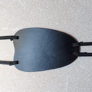 May include: A black leather slingshot pouch with attached black elastic bands. The pouch is oval-shaped with a textured surface. The elastic bands are secured to the pouch with small metal grommets.