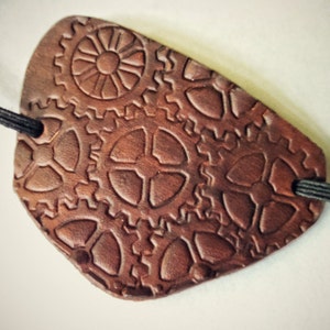 May include: A brown leather eye patch with a steampunk design featuring embossed gears. The patch is attached to a black elastic band.