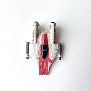 May include: A white and red toy spaceship with black details. The spaceship has two wings and a pointed nose.