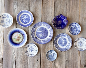 May include: A collection of vintage blue and white decorative plates of various sizes and designs, including floral, landscape, and classical scenes. The plates are arranged on a rustic wooden surface.
