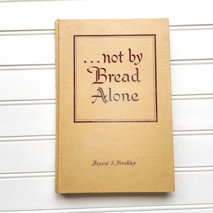 May include: A tan hardcover book with the title "...not by Bread Alone" in brown lettering. The author, Bryant S. Hinckley, is printed on the bottom of the cover.