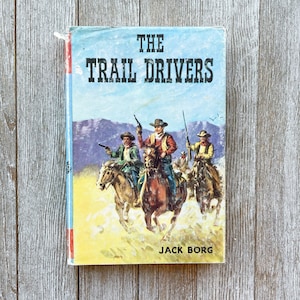 May include: A vintage paperback book cover with a blue background and a scene of cowboys on horseback. The title of the book is "The Trail Drivers" by Jack Borg.