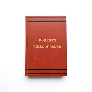 May include: A small, rectangular book with a textured, reddish-brown cover. The title "ROBERT'S RULES OF ORDER" is printed in gold lettering. Black lines border the top and bottom of the book.