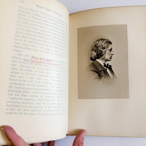 May include: An open book with text and a sepia-toned portrait of a man in a suit and bow tie. The portrait is on the right page, while the left page has printed text. The book is held open by a hand.