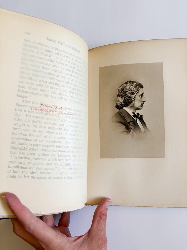 May include: An open book with text and a sepia-toned portrait of a man in a suit and bow tie. The portrait is on the right page, while the left page has printed text. The book is held open by a hand.