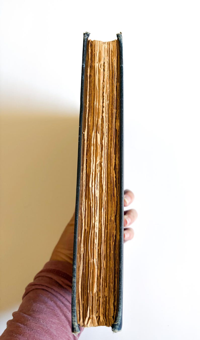 May include: A thick, antique book with aged, yellowed pages and a dark blue cover. The book's spine is visible, revealing the worn edges of the pages. The book is held by a person, with a white background.