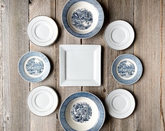 May include: A set of eight blue and white ceramic plates with a gold trim. The plates feature a blue and white floral pattern and a scene of a house in the countryside. The plates are arranged in a circle on a wooden surface.