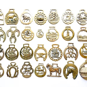 May include: A collection of twenty-eight brass horse brasses featuring various embossed designs including animals, ships, buildings, and symbols. Each piece has a loop at the top for attachment, showcasing a metallic gold finish and intricate details.