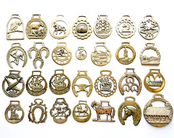 May include: A collection of twenty-eight brass horse brasses featuring various embossed designs including animals, ships, buildings, and symbols. Each piece has a loop at the top for attachment, showcasing a metallic gold finish and intricate details.