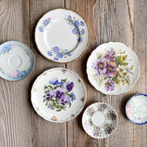 May include: Six decorative plates with floral designs. The plates feature various floral patterns with blue, purple, and white flowers. The plates are arranged on a wooden surface.