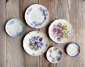 May include: Six decorative plates with floral designs. The plates feature various floral patterns with blue, purple, and white flowers. The plates are arranged on a wooden surface.