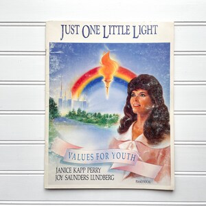 May include: A book cover with a rainbow and a torch, featuring the title "Just One Little Light" and the subtitle "Values for Youth". The authors are Janice Kapp Perry and Joy Saunders Lundberg. The cover also includes a woman with long brown hair.