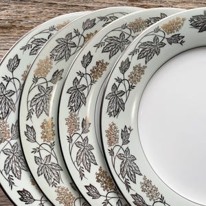 May include: A set of four white ceramic plates with a black and gold leaf and floral design around the rim. The plates have a black border and are arranged on a wooden surface.