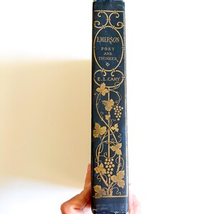 May include: A vintage book spine with the title "Emerson Poet and Thinker" in gold lettering on a dark blue background. The spine features decorative gold floral and grape vine designs. The author's name, E.L. Cary, is also in gold.