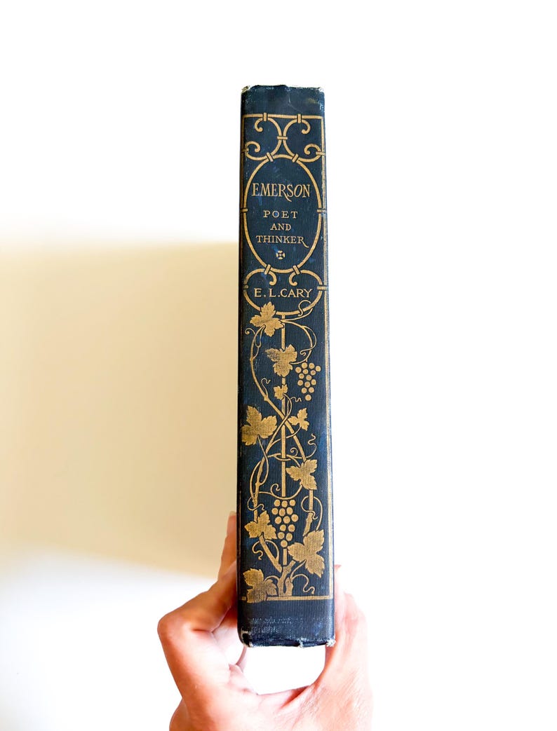 May include: A vintage book spine with the title "Emerson Poet and Thinker" in gold lettering on a dark blue background. The spine features decorative gold floral and grape vine designs. The author's name, E.L. Cary, is also in gold.