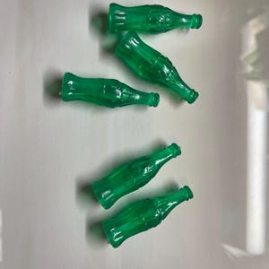 May include: Six transparent green miniature soda bottles. The bottles are arranged in a scattered pattern. The bottles have a classic soda bottle shape with a narrow neck and a wider base. The bottles are made of a shiny, translucent material.