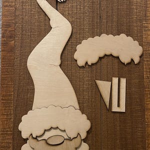 May include: Wooden gnome craft pieces. The set includes a long, curved hat with a bell, a cloud-shaped beard, and two additional pieces. The pieces are made of unfinished wood, ready for painting or decorating. Perfect for DIY holiday crafts.