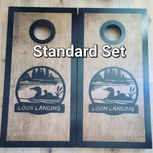 May include: A pair of rectangular wooden cornhole boards with black frames and a natural wood grain surface. Each board features a circular hole at the top and a black graphic of a loon on a lake with the text "LOON LANDING" and "Standard Set".