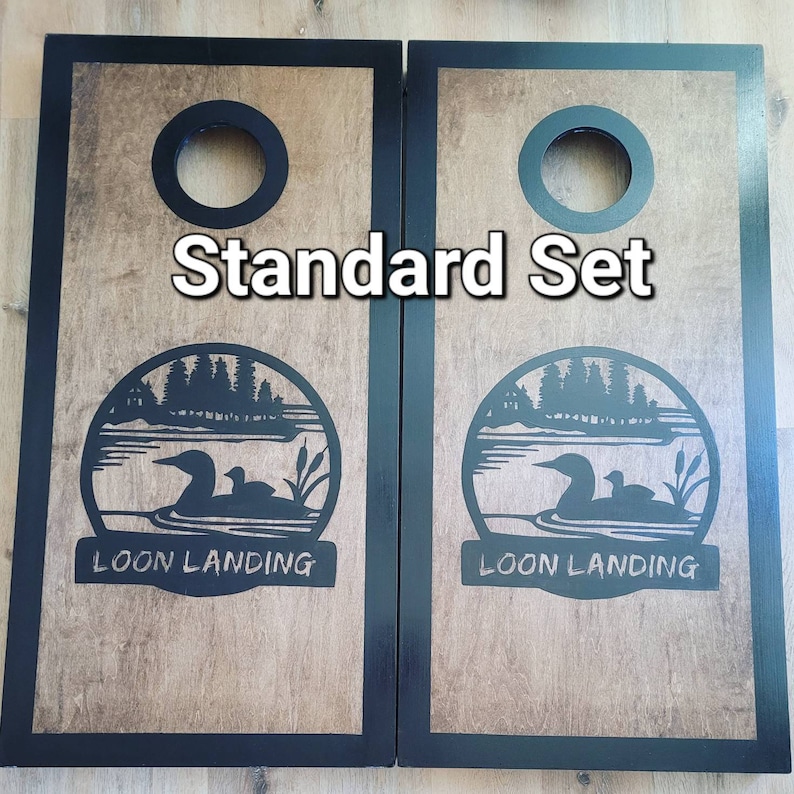 May include: A pair of rectangular wooden cornhole boards with black frames and a natural wood grain surface. Each board features a circular hole at the top and a black graphic of a loon on a lake with the text "LOON LANDING" and "Standard Set".
