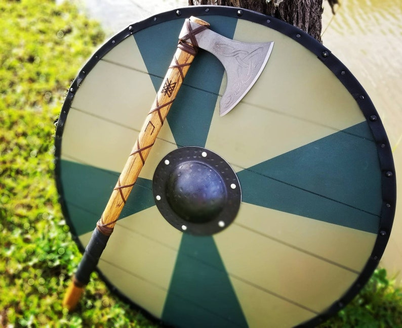 May include: A Viking-style shield with a green and tan geometric design, featuring a black metal boss and rim. A wooden-handled axe with a silver blade rests on the shield. The axe handle is wrapped with brown leather.